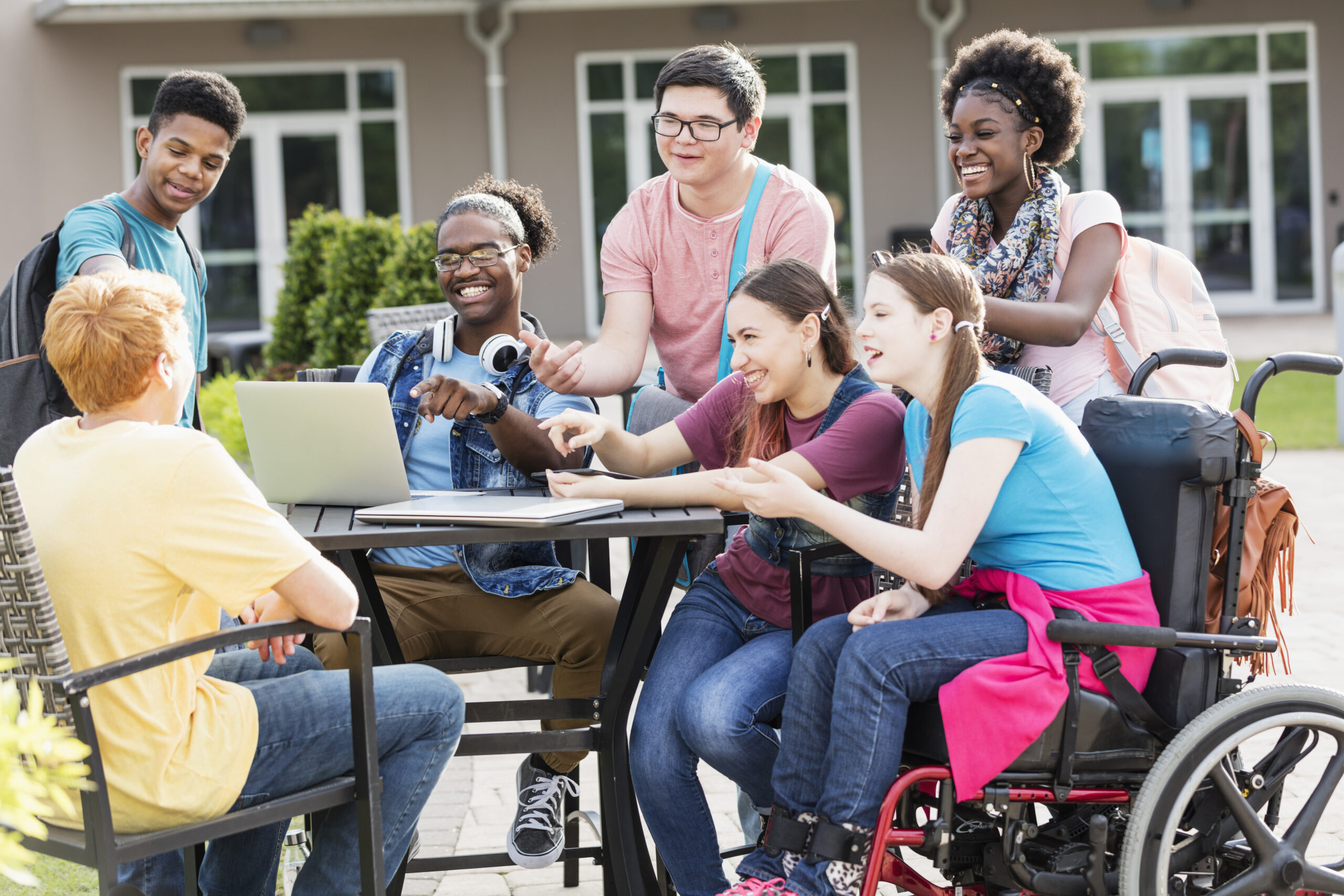 A diverse group of teens, including a student in a wheelchair, gathered around an outdoor table looking at a laptop together.